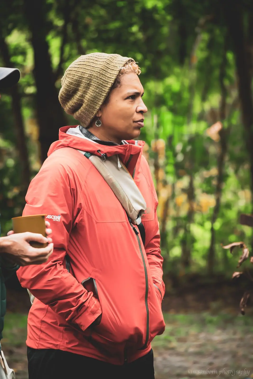 A person in a knitted beige hat and coral rain jacket stands among bright green trees, holding a cup, looking off to one side.