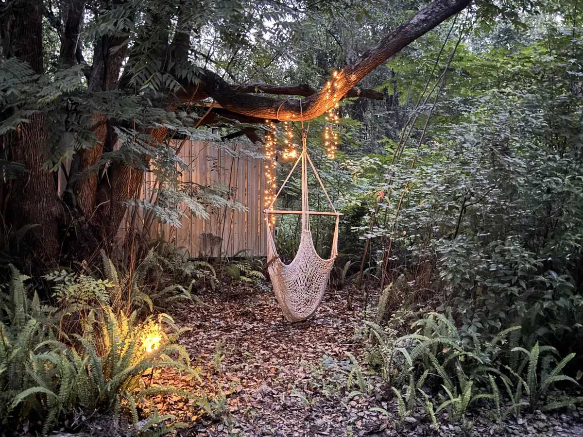 A white rope hammock chair hanging from a long oak limb wrapped in fairy lights, over a leaf-covered clearing ringed by ferns and dense green forest at dusk.