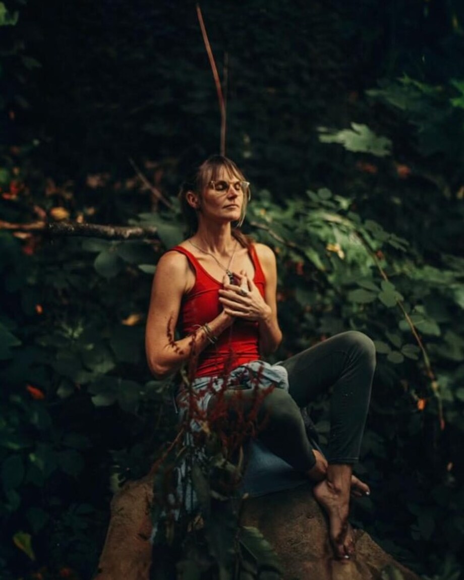 Summer Crider seated barefoot on a fallen log deep in green undergrowth, hands folded over their heart, eyes closed and face turned toward the light.